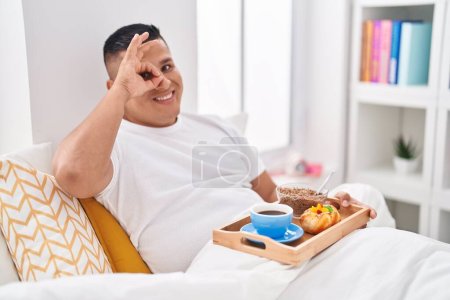 Téléchargez les photos : Jeune homme hispanique manger le petit déjeuner dans le lit souriant heureux faire ok signe avec la main sur les yeux en regardant à travers les doigts - en image libre de droit