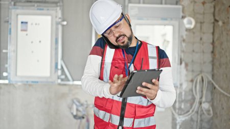 Photo for Young latin man builder talking on smartphone using touchpad at construction site - Royalty Free Image