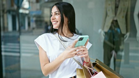 Foto de Mujer hispana hermosa joven usando teléfono inteligente sosteniendo bolsas de compras en la tienda de ropa - Imagen libre de derechos