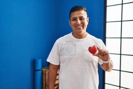 Photo for Hispanic young man holding red heart at gym looking positive and happy standing and smiling with a confident smile showing teeth - Royalty Free Image
