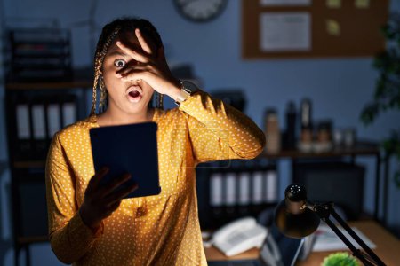 Foto de Mujer afroamericana con trenzas trabajando en la oficina por la noche con tableta asomándose en shock cubriendo la cara y los ojos con la mano, mirando a través de los dedos con expresión avergonzada. - Imagen libre de derechos