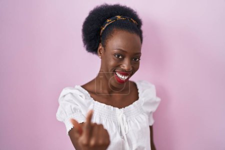 Foto de Mujer africana con el pelo rizado de pie sobre fondo rosa señas vienen aquí gesto con la mano invitando a la bienvenida feliz y sonriente - Imagen libre de derechos