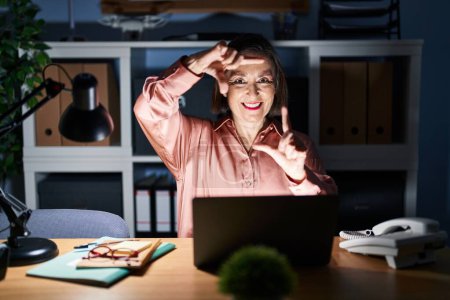 Téléchargez les photos : Femme hispanique du Moyen Age travaillant à l'aide d'un ordinateur portable tard dans la nuit souriant faire cadre avec les mains et les doigts avec le visage heureux. concept de créativité et photographie. - en image libre de droit