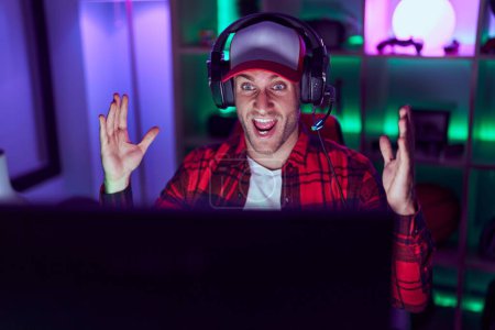 Téléchargez les photos : Jeune homme caucasien jouant à des jeux vidéo célébrant la victoire avec un sourire heureux et l'expression gagnante avec les mains levées - en image libre de droit