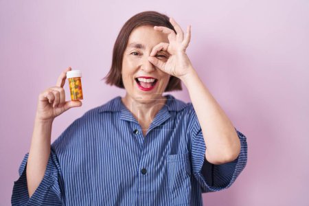 Téléchargez les photos : Moyen Age hispanique femme tenant des pilules souriant heureux faire ok signe avec la main sur les yeux en regardant à travers les doigts - en image libre de droit