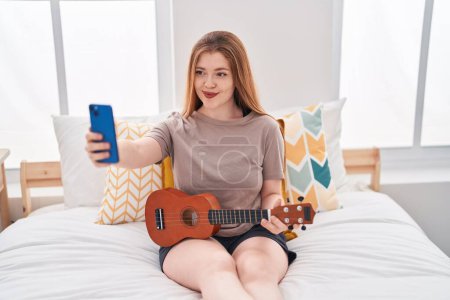 Téléchargez les photos : Jeune rousse femme faire selfie par smarthone tenant ukulele dans la chambre - en image libre de droit