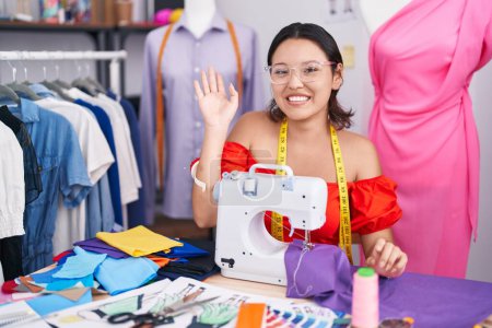 Téléchargez les photos : Jeune femme hispanique couturière designer utilisant une machine à coudre renonçant à dire bonjour heureux et souriant, geste de bienvenue convivial - en image libre de droit