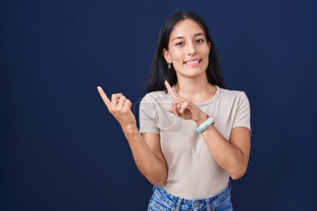 Photo for Young hispanic woman standing over blue background smiling and looking at the camera pointing with two hands and fingers to the side. - Royalty Free Image