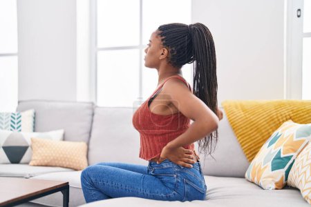 Téléchargez les photos : Femme afro-américaine souffrant de maux de dos assis sur le canapé à la maison - en image libre de droit