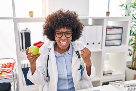 Photo for Black doctor woman with curly hair holding green apple screaming proud, celebrating victory and success very excited with raised arms - Royalty Free Image