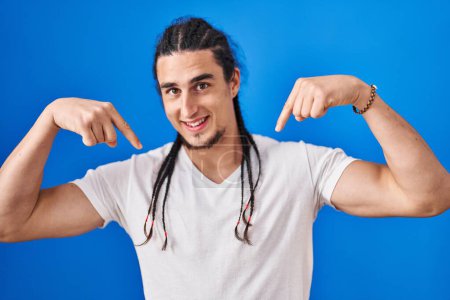 Photo for Hispanic man with long hair standing over blue background looking confident with smile on face, pointing oneself with fingers proud and happy. - Royalty Free Image
