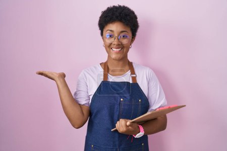 Téléchargez les photos : Jeune femme afro-américaine portant tablier de serveuse professionnel tenant presse-papiers pointant de côté avec les mains ouvertes paumes montrant l'espace de copie, présentant la publicité souriant heureux excité - en image libre de droit
