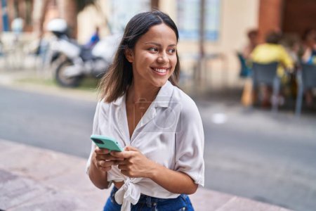 Téléchargez les photos : Jeune femme hispanique souriant confiant en utilisant smartphone à la rue - en image libre de droit
