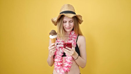 Téléchargez les photos : Jeune femme blonde touriste tenant crème glacée en utilisant smartphone sur fond jaune isolé - en image libre de droit