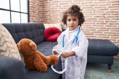 Photo for Adorable hispanic girl wearing doctor uniform examining teddy bear at home - Royalty Free Image