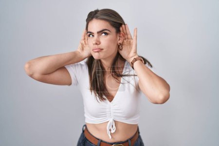 Foto de Joven hermosa mujer con camiseta blanca casual tratando de escuchar ambas manos en el gesto de la oreja, curioso por chismes. problema auditivo, sordo - Imagen libre de derechos
