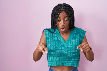 Téléchargez les photos : Jeune afro-américain avec des tresses debout sur fond rose pointant vers le bas avec les doigts montrant la publicité, visage surpris et bouche ouverte - en image libre de droit