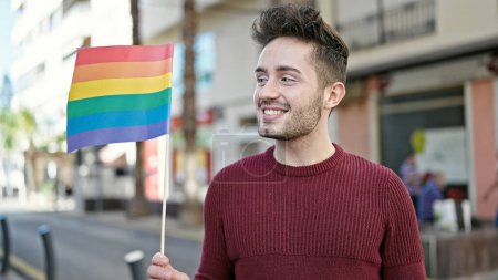 Téléchargez les photos : Jeune homme hispanique souriant confiant tenant drapeau arc-en-ciel dans la rue - en image libre de droit