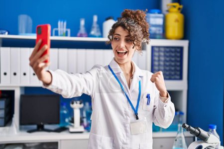 Foto de Mujer hispana con el pelo rizado trabajando en laboratorio científico haciendo selfie gritando orgullosa, celebrando la victoria y el éxito muy emocionada con el brazo levantado - Imagen libre de derechos