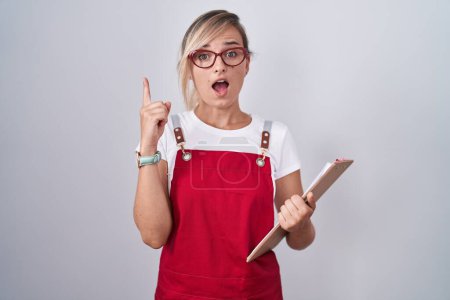 Photo for Young blonde woman wearing waiter uniform holding clipboard pointing finger up with successful idea. exited and happy. number one. - Royalty Free Image