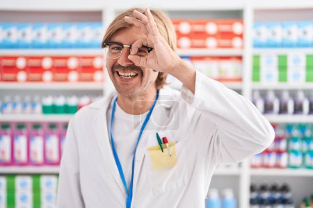 Foto de Hombre caucásico con bigote trabajando en farmacia haciendo buen gesto con la mano sonriente, los ojos mirando a través de los dedos con la cara feliz. - Imagen libre de derechos