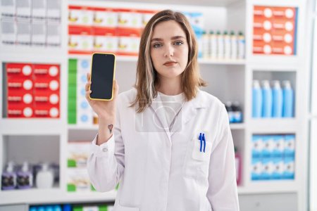 Photo for Blonde caucasian woman working at pharmacy drugstore showing smartphone screen thinking attitude and sober expression looking self confident - Royalty Free Image