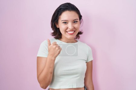 Foto de Mujer joven hispana de pie sobre fondo rosa haciendo un gesto feliz pulgares hacia arriba con la mano. aprobando la expresión mirando a la cámara mostrando éxito. - Imagen libre de derechos