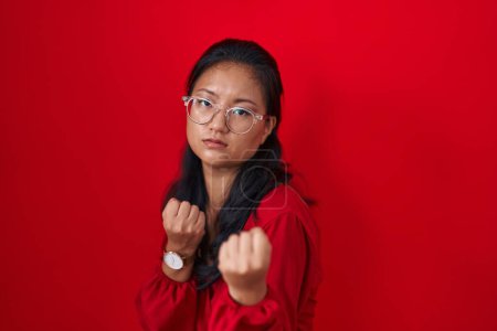 Téléchargez les photos : Asiatique jeune femme debout sur fond rouge prêt à se battre avec poing geste de défense, visage en colère et bouleversé, peur du problème - en image libre de droit