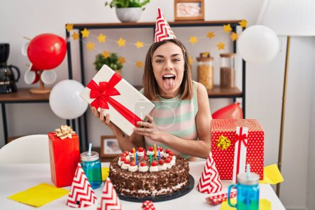 Photo for Hispanic woman celebrating birthday with cake holding gift sticking tongue out happy with funny expression. - Royalty Free Image