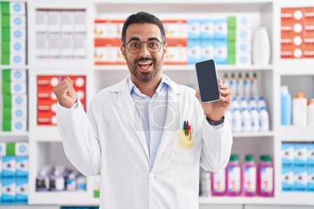 Téléchargez les photos : Homme hispanique avec barbe travaillant à la pharmacie pharmacie montrant écran smartphone criant fier, célébrant la victoire et le succès très excité avec le bras levé - en image libre de droit