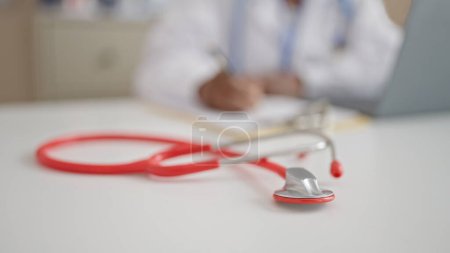 Photo for African american woman doctor using laptop writing on clipboard at clinic - Royalty Free Image