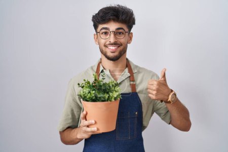 Téléchargez les photos : Homme arabe avec barbe tenant pot de plante verte faisant des pouces heureux geste avec la main. approuvant l'expression regardant la caméra montrant le succès. - en image libre de droit