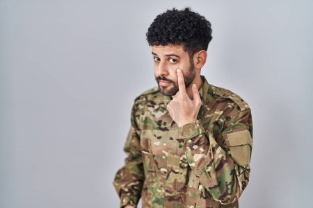 Foto de Hombre árabe vistiendo uniforme del ejército de camuflaje apuntando al ojo observándote gesto, expresión sospechosa - Imagen libre de derechos