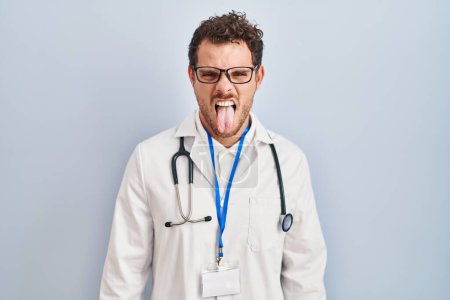 Foto de Joven hombre hispano vistiendo uniforme médico y estetoscopio sacando la lengua feliz con expresión divertida. concepto de emoción. - Imagen libre de derechos