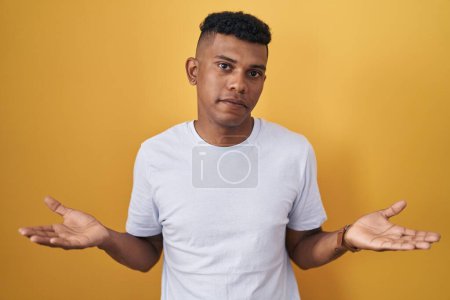 Téléchargez les photos : Jeune homme hispanique debout sur fond jaune désemparé et confondu avec les bras ouverts, aucune idée concept. - en image libre de droit