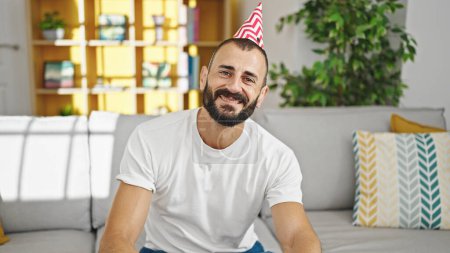 Foto de Joven hombre hispano sonriendo confiado usando sombrero de cumpleaños en casa - Imagen libre de derechos