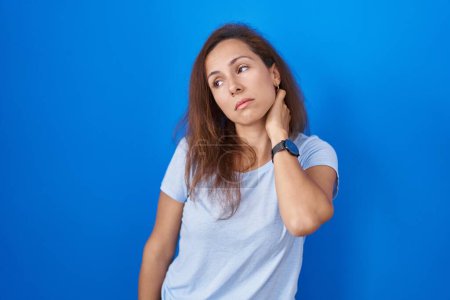 Téléchargez les photos : Femme brune debout sur fond bleu souffrant d'une blessure au cou, toucher le cou avec la main, douleur musculaire - en image libre de droit
