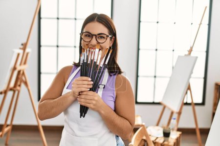 Téléchargez les photos : Jeune belle artiste hispanique souriante confiante tenant pinceaux au studio d'art - en image libre de droit