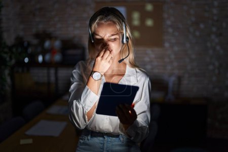 Téléchargez les photos : Jeune femme blonde travaillant au bureau la nuit sentant quelque chose de puant et dégoûtant, odeur intolérable, retenant la respiration avec les doigts sur le nez. mauvaise odeur - en image libre de droit