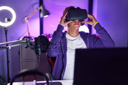 Téléchargez les photos : Jeune adolescent hispanique streamer jouer à un jeu vidéo en utilisant des lunettes de réalité virtuelle à la salle de jeu - en image libre de droit