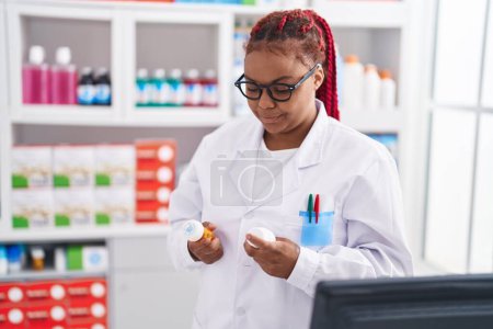 Téléchargez les photos : Femme afro-américaine pharmacien tenant des bouteilles de pilules à la pharmacie - en image libre de droit