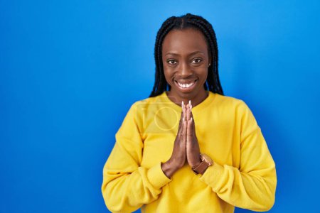 Photo for Beautiful black woman standing over blue background praying with hands together asking for forgiveness smiling confident. - Royalty Free Image