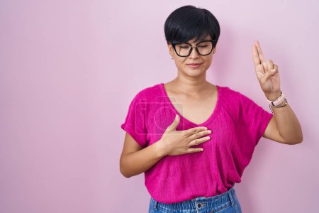 Foto de Joven mujer asiática con el pelo corto de pie sobre fondo rosa sonriendo jurando con la mano en el pecho y los dedos hacia arriba, haciendo una promesa de lealtad juramento - Imagen libre de derechos