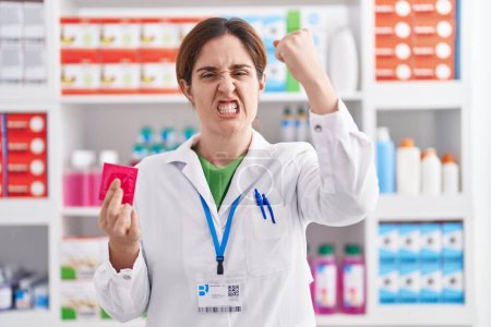 Téléchargez les photos : Femme brune travaillant à la pharmacie pharmacie tenant préservatif ennuyé et frustré criant de colère, criant fou de colère et la main levée - en image libre de droit