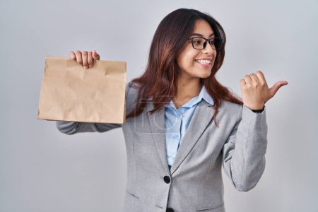 Photo for Hispanic young woman holding take away bag pointing thumb up to the side smiling happy with open mouth - Royalty Free Image