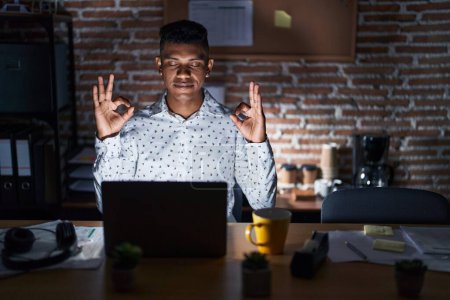 Téléchargez les photos : Jeune homme hispanique travaillant au bureau la nuit se détendre et sourire les yeux fermés en faisant geste de méditation avec les doigts. concept de yoga. - en image libre de droit