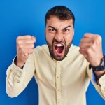 Handsome hispanic man standing over blue background angry and mad raising fists frustrated and furious while shouting with anger. rage and aggressive concept.