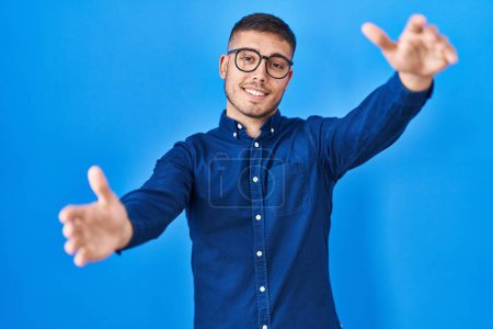 Téléchargez les photos : Jeune homme hispanique portant des lunettes sur fond bleu regardant la caméra souriant à bras ouverts pour un câlin. expression joyeuse embrassant le bonheur. - en image libre de droit