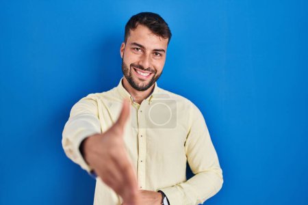 Téléchargez les photos : Bel homme hispanique debout sur fond bleu souriant amical offrant poignée de main comme salutation et accueil. entreprise prospère. - en image libre de droit