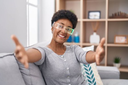 Téléchargez les photos : Jeune femme afro-américaine portant des lunettes de réalité virtuelle assise sur le canapé regardant la caméra souriant à bras ouverts pour se faire câliner. expression joyeuse embrassant le bonheur. - en image libre de droit